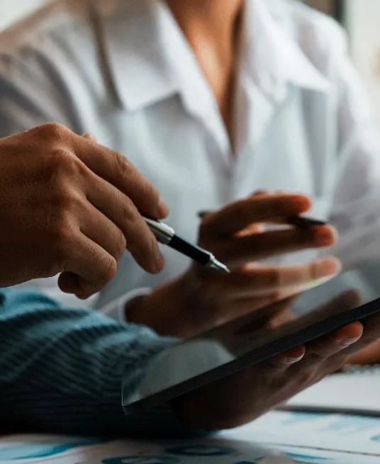 Two workers viewing a tablet. There are notebooks and computers displaying graphs on the table they are working on.
