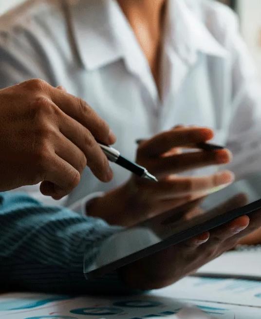 Two workers viewing a tablet. There are notebooks and computers displaying graphs on the table they are working on.