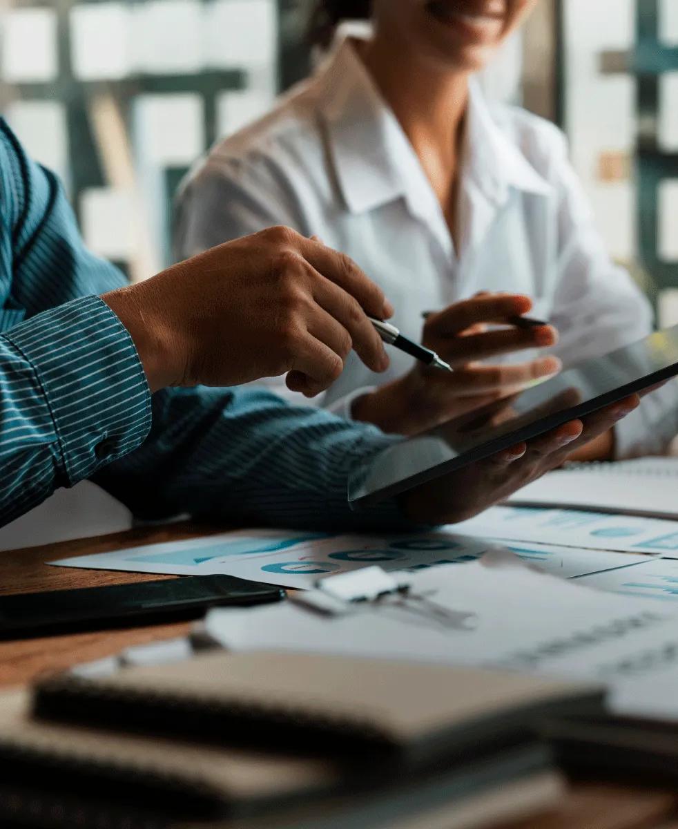 Two workers viewing a tablet. There are notebooks and computers displaying graphs on the table they are working on.