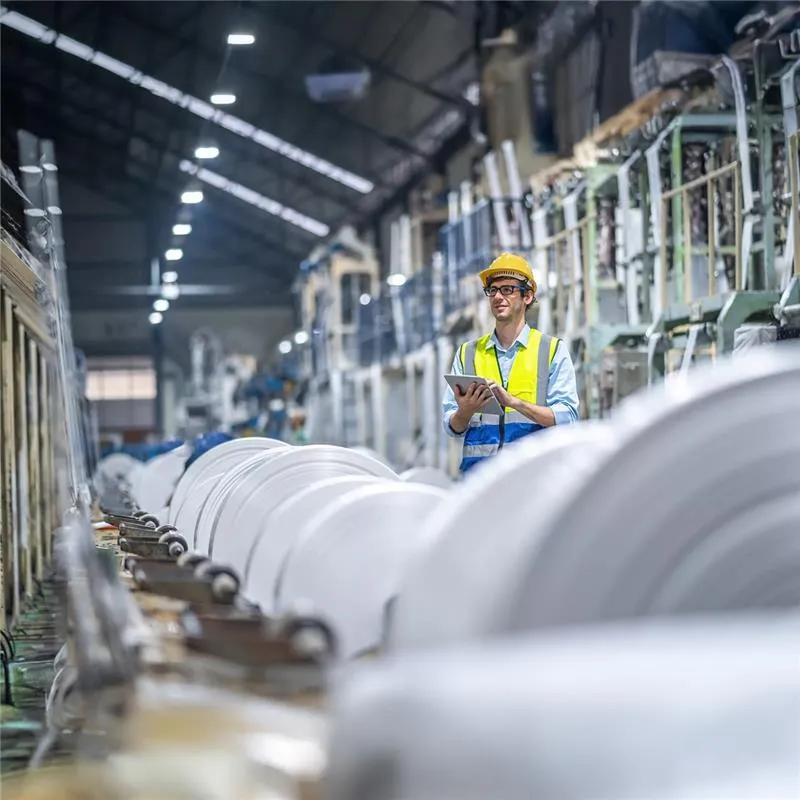 A factory worker wearing a yellow safety vest and hard hat stands in an aisle, looking over a tablet. The foreground and background are dominated by large, white rolls of material stacked high in the industrial storage area.