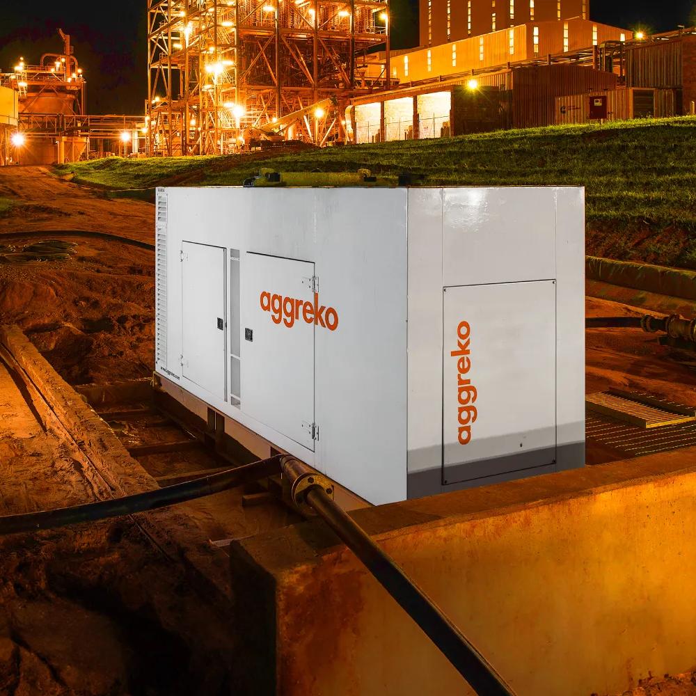A night scene featuring an industrial container unit with "Aggreko" branding, surrounded by illuminated infrastructure and wet ground.