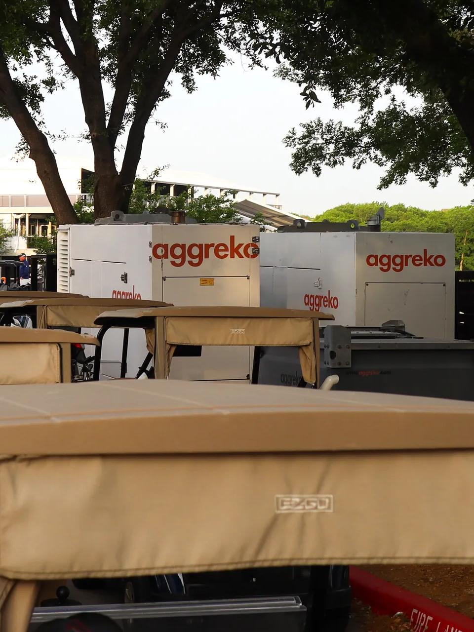 Several golf carts in the shade with two white Aggreko units.