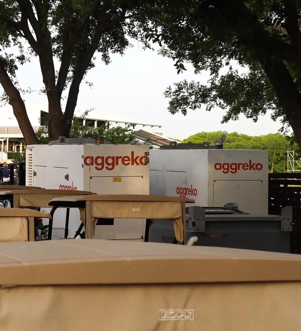 Several golf carts in the shade with two white Aggreko units.