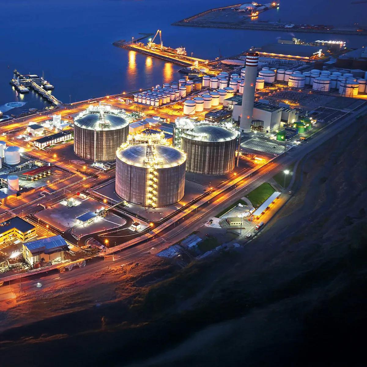 An aerial view of an industrial port at night, featuring illuminated storage tanks and shipping facilities along the water.