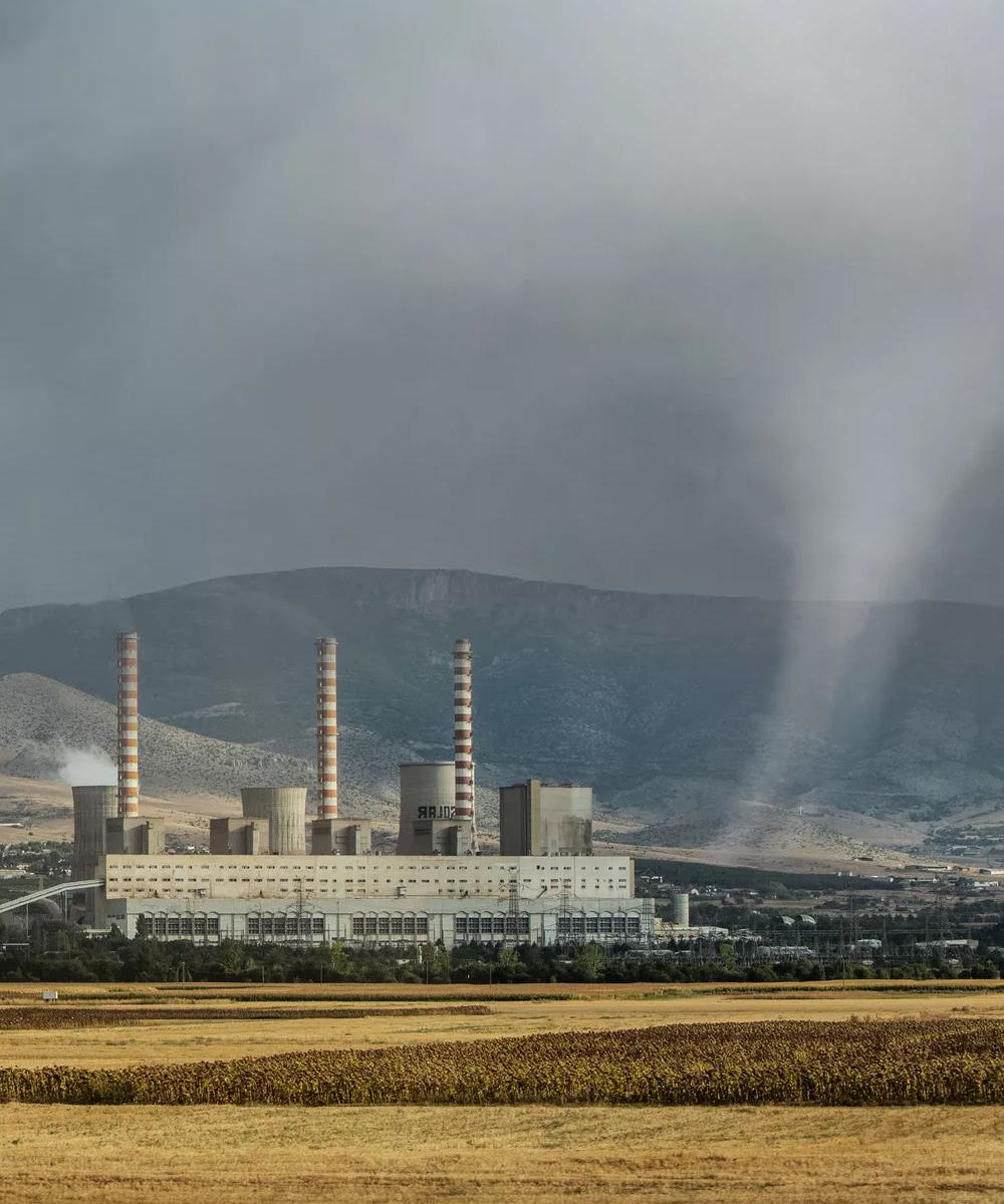 A power plant with cooling towers and smokestacks, set against a backdrop of mountains and cloudy skies, with fields in the foreground.