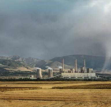 A power plant with cooling towers and smokestacks, set against a backdrop of mountains and cloudy skies, with fields in the foreground.