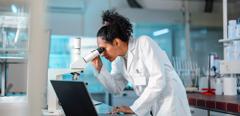 Young Hispanic scientist wearing a lab coat, looking under microscope while using laptop in a laboratory.