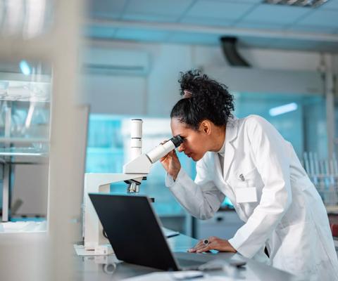 Young Hispanic scientist wearing a lab coat, looking under microscope while using laptop in a laboratory.