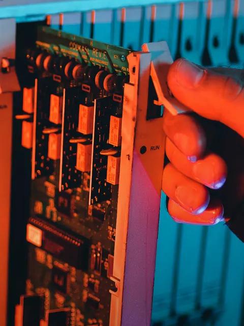 A close-up of a person's hand, lit brightly in red, adjusting a switch on an industrial circuit board. The circuit board features relays and various components.