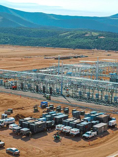 Aerial view of a variety of containerized industrial equipment and generators staged on a gravel lot near a green field with several large wind turbines in the background.