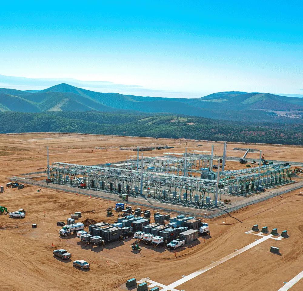 Aerial view of a variety of containerized industrial equipment and generators staged on a gravel lot near a green field with several large wind turbines in the background.