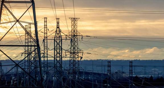Silhouetted power lines and towers against a sunset sky, with soft clouds and distant hills in the background.