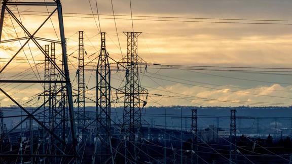 Silhouetted power lines and towers against a sunset sky, with soft clouds and distant hills in the background.