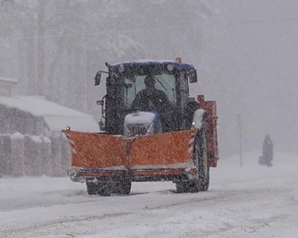 A front view of a snow plow pushing through heavy snowfall, with the driver visible inside.