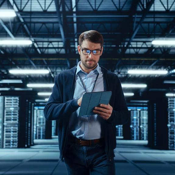 Male IT Specialist Walks Between Row of Operational Server Racks in Data Center. Engineer Uses Tablet Computer for Maintenance. Concept for Cloud Computing, Artificial Intelligence, Cybersecurity.