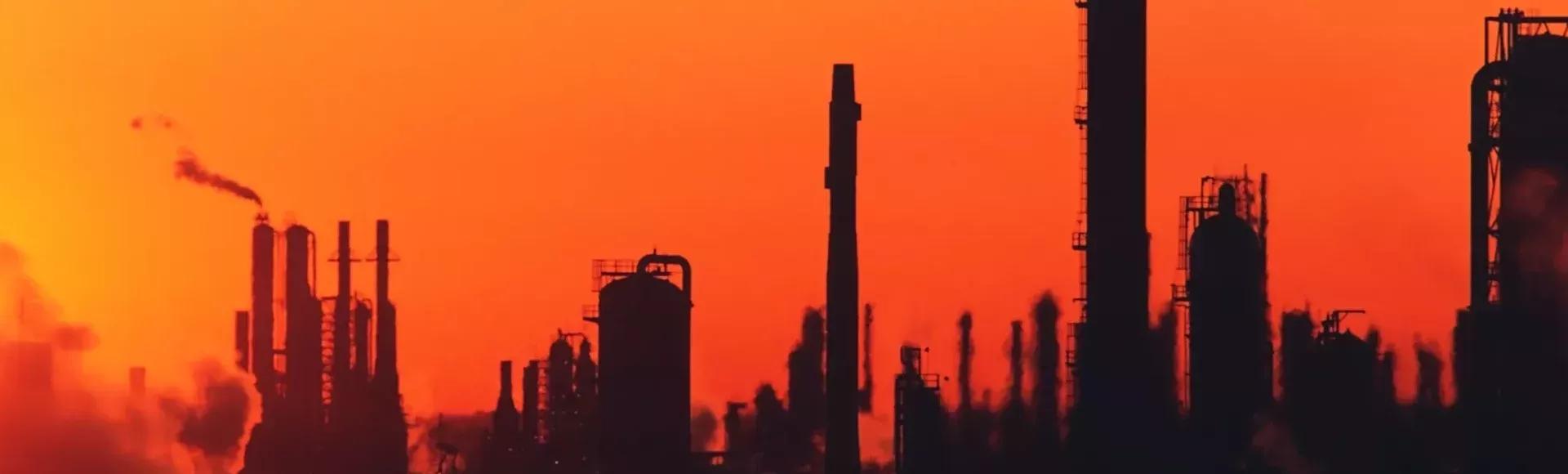 The silhouette of an industrial plant with smokestacks and pipes against a vibrant orange and red sunset.