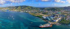 Aerial view of a harbor with a container ship docked, surrounded by green hills, buildings, and scattered sailboats in clear blue water under cloudy skies.