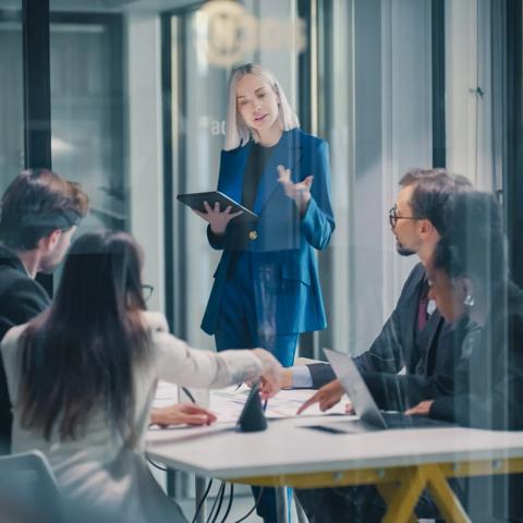 Businesswoman Leads a Strategy Meeting in a Glass Office, Using a Digital Tablet to Explain Goals While a Corporate Team Reviews Data, Negotiates Priorities, Aligns Projects