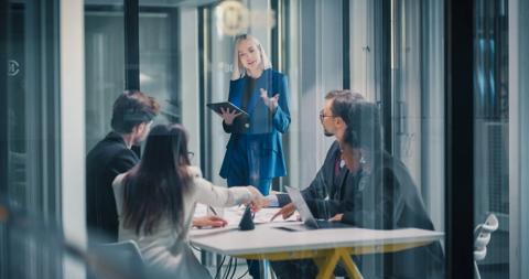 Businesswoman Leads a Strategy Meeting in a Glass Office, Using a Digital Tablet to Explain Goals While a Corporate Team Reviews Data, Negotiates Priorities, Aligns Projects