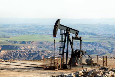 An oil pump jack operates on a hillside, overlooking green fields and rugged terrain in the background.