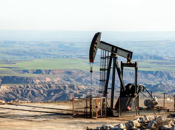 An oil pump jack operates on a hillside, overlooking green fields and rugged terrain in the background.