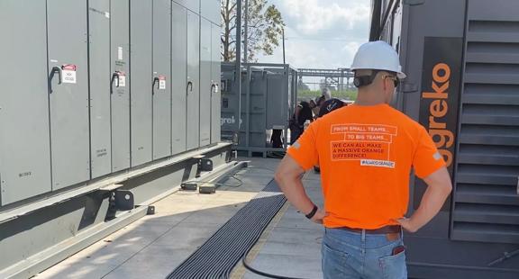 Travailleur portant une chemise orange Aggreko et un casque de sécurité blanc, debout à côté d’un équipement électrique industriel dans une installation extérieure