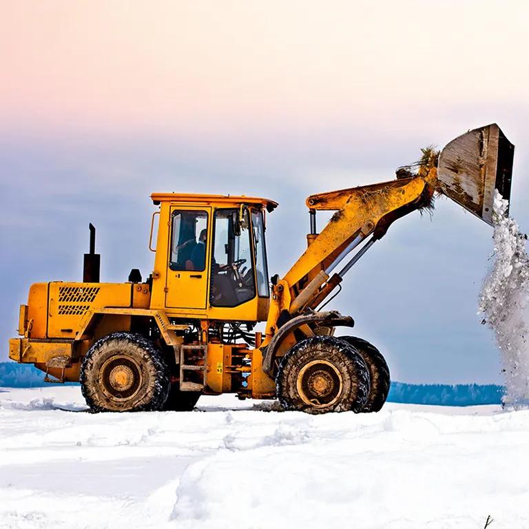 A bright yellow front loader is scooping a load of white material, likely snow or a byproduct, in a sunny, snowy or icy industrial environment.
