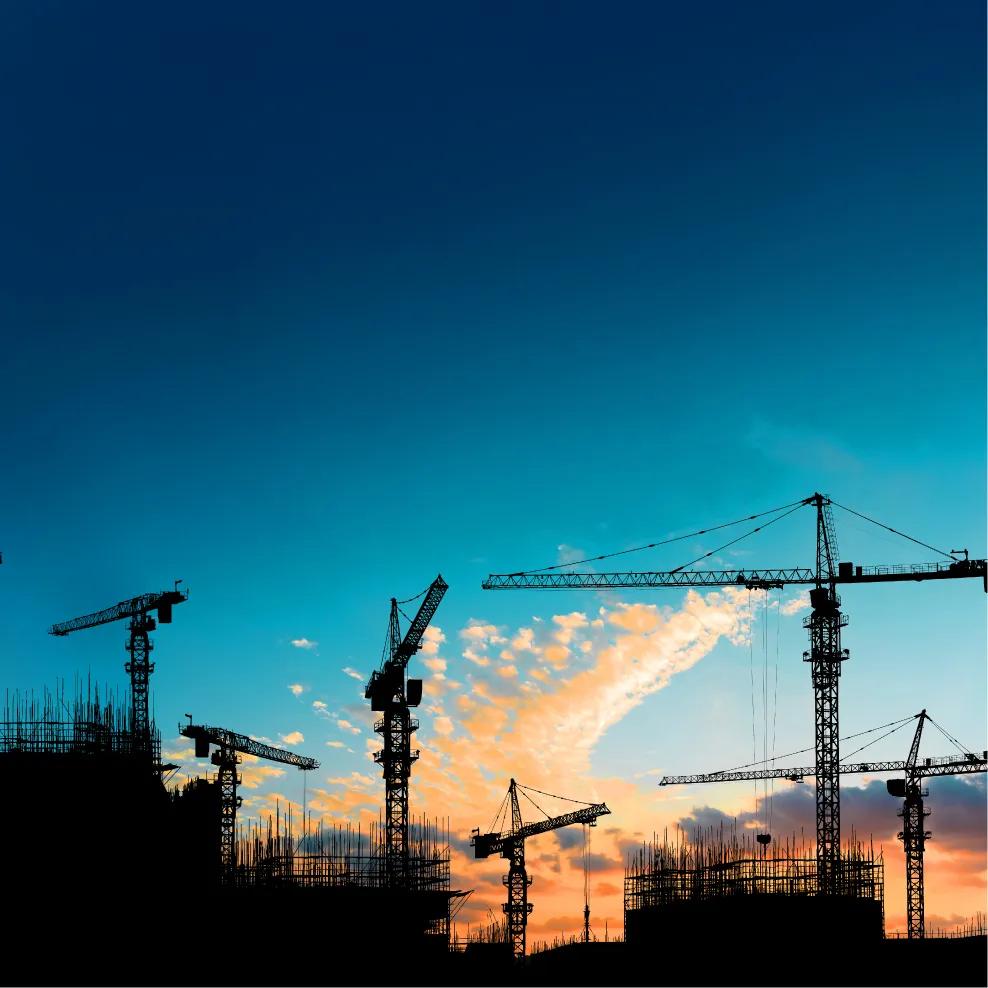 Les silhouettes de deux grues de chantier et d’une ossature de bâtiment se détachent sur un ciel bleu foncé au coucher du soleil.
