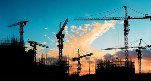 Six tower cranes at a worksite next to a building in process against a sunset sky