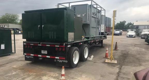 The rear view of a black dual-axle flatbed trailer, carrying a large dark green industrial enclosure and a gray, modular unit. The trailer is parked on an asphalt or concrete lot with orange traffic cones nearby.