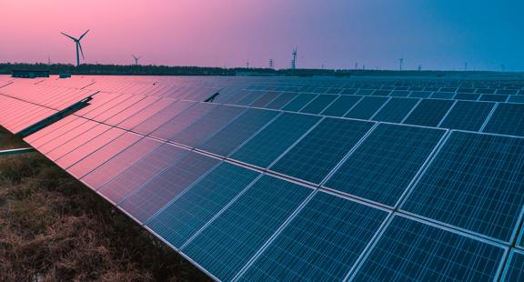 Un champ de panneaux solaires sous un ciel coloré, avec des éoliennes visibles en arrière-plan.