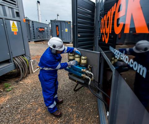 A worker in a blue uniform and helmet operates equipment near containers with the logo Aggreko in an outdoor setting.