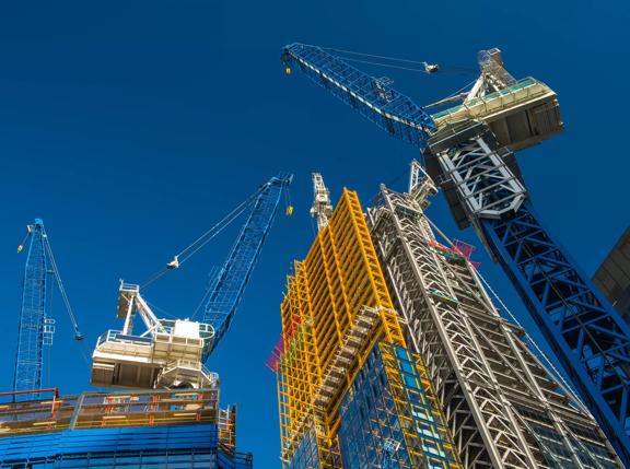 Construction site with multiple cranes and a partially completed high-rise building against a clear blue sky.