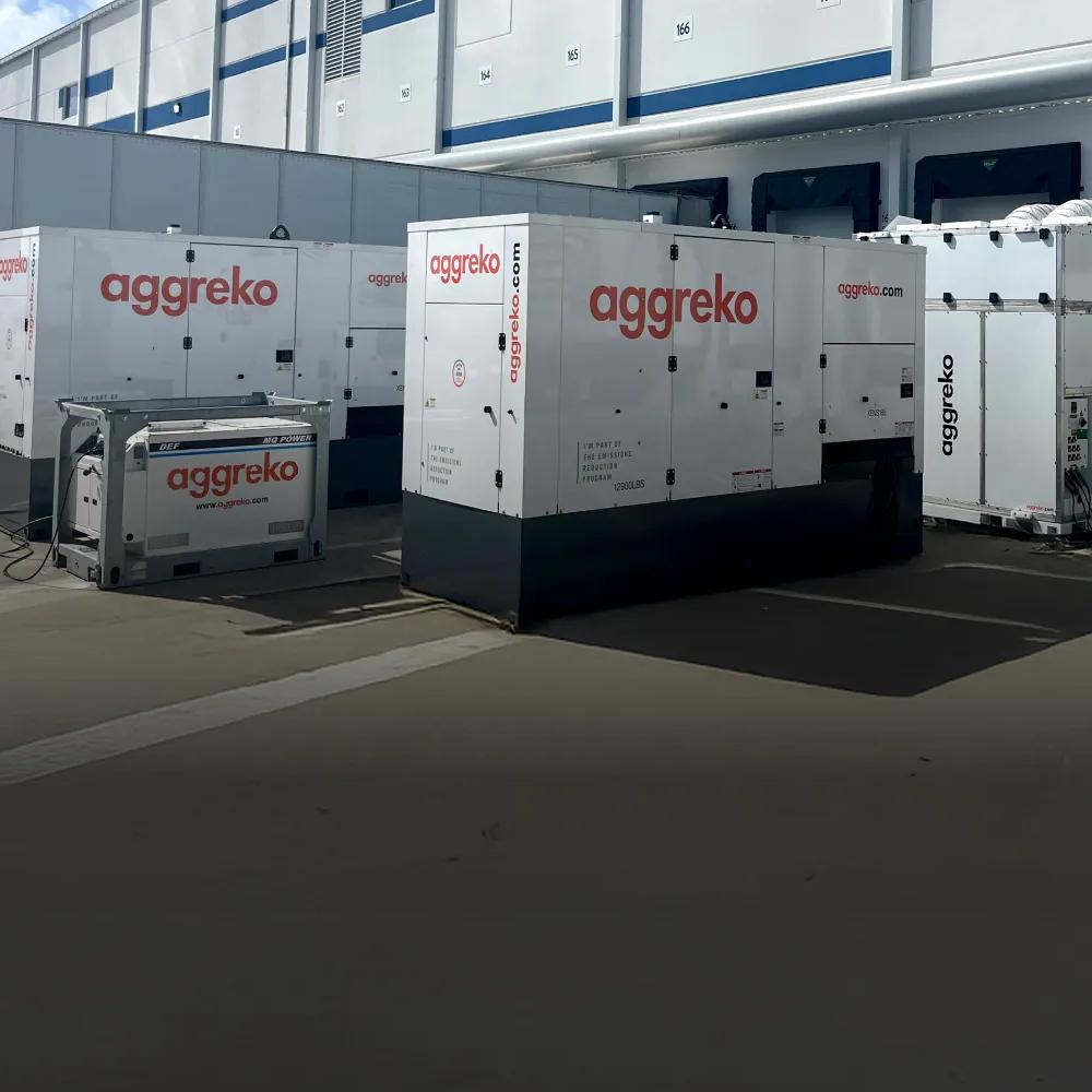 Two white Aggreko generators and a fuel tank on a buildings parking lot next to a truck