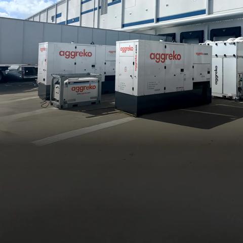 Two white Aggreko generators and a fuel tank on a buildings parking lot next to a truck