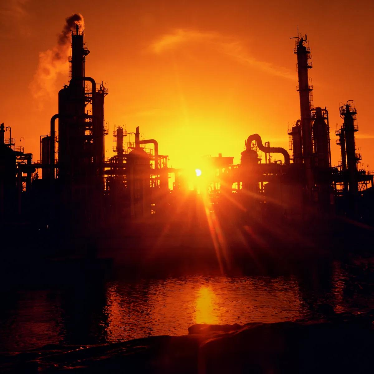 Silhouette of industrial structures against a vibrant orange sunset, with smoke rising from the facilities.