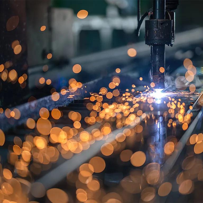 A close-up, dark image of a welding or cutting operation, showing a shower of bright orange sparks against a blurry industrial background.