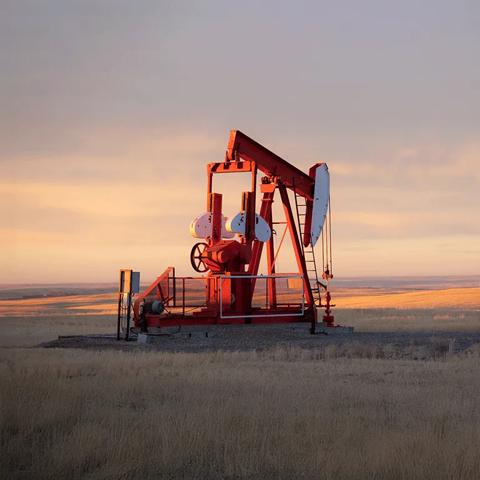 A large, red oil pumpjack is prominently featured on the right, silhouetted against a vast, open landscape under a dramatic golden and orange sunset.