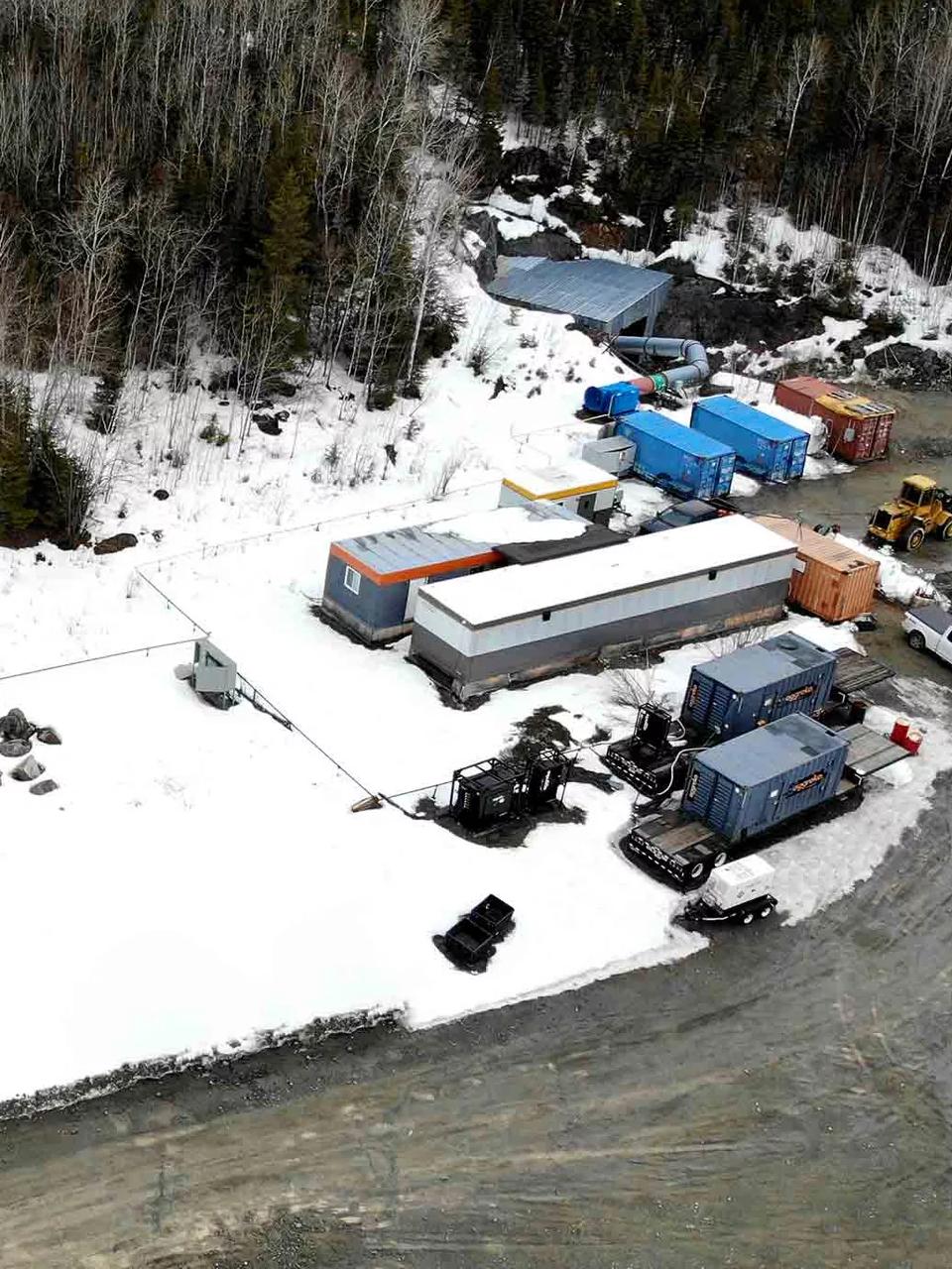 An aerial view of a mining site with several buildings and vehicles in a snowy, mountainous landscape. 