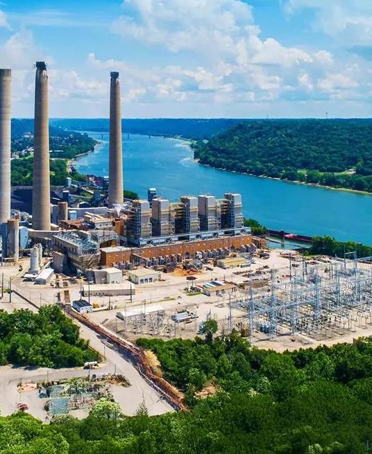 An aerial view of a large power plant and associated electrical substation next to a wide river and forested hills.