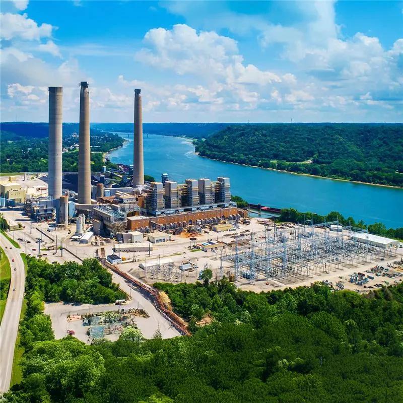 An aerial view of a large power plant and associated electrical substation next to a wide river and forested hills.