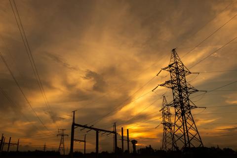 Black outline of a transmission tower at the front and multiple transmission towers in the background, against a cloudy grey and yellow sky.