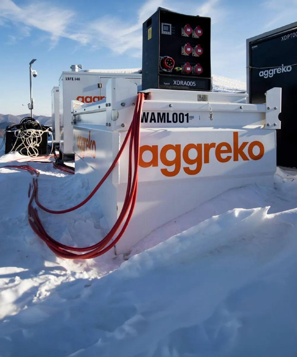 Vue panoramique d’un réservoir de carburant Aggreko blanc avec le logo orange et d’autres équipements Aggreko au sommet d’une montagne enneigée avec des arbres