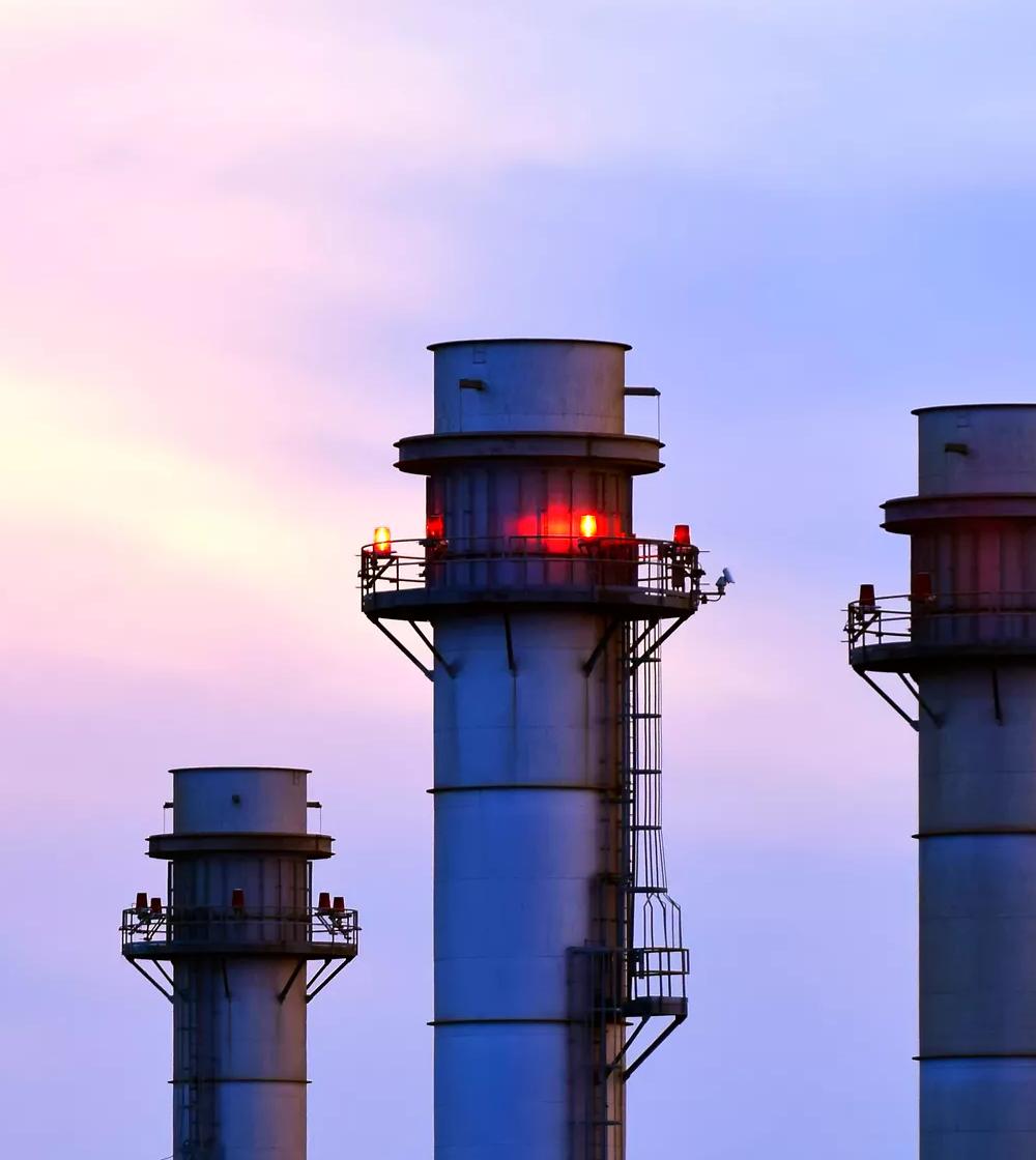 Night-view of a lit-up refinery at a distance.  View of tall industrial smokestacks with red safety lights at dusk.