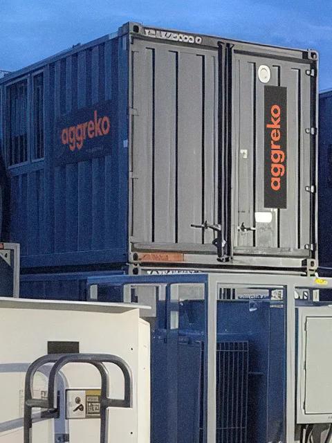Multiple gray, Aggreko industrial units stacked on one another. A worker in uniform is flipping panel switches beside the units.  