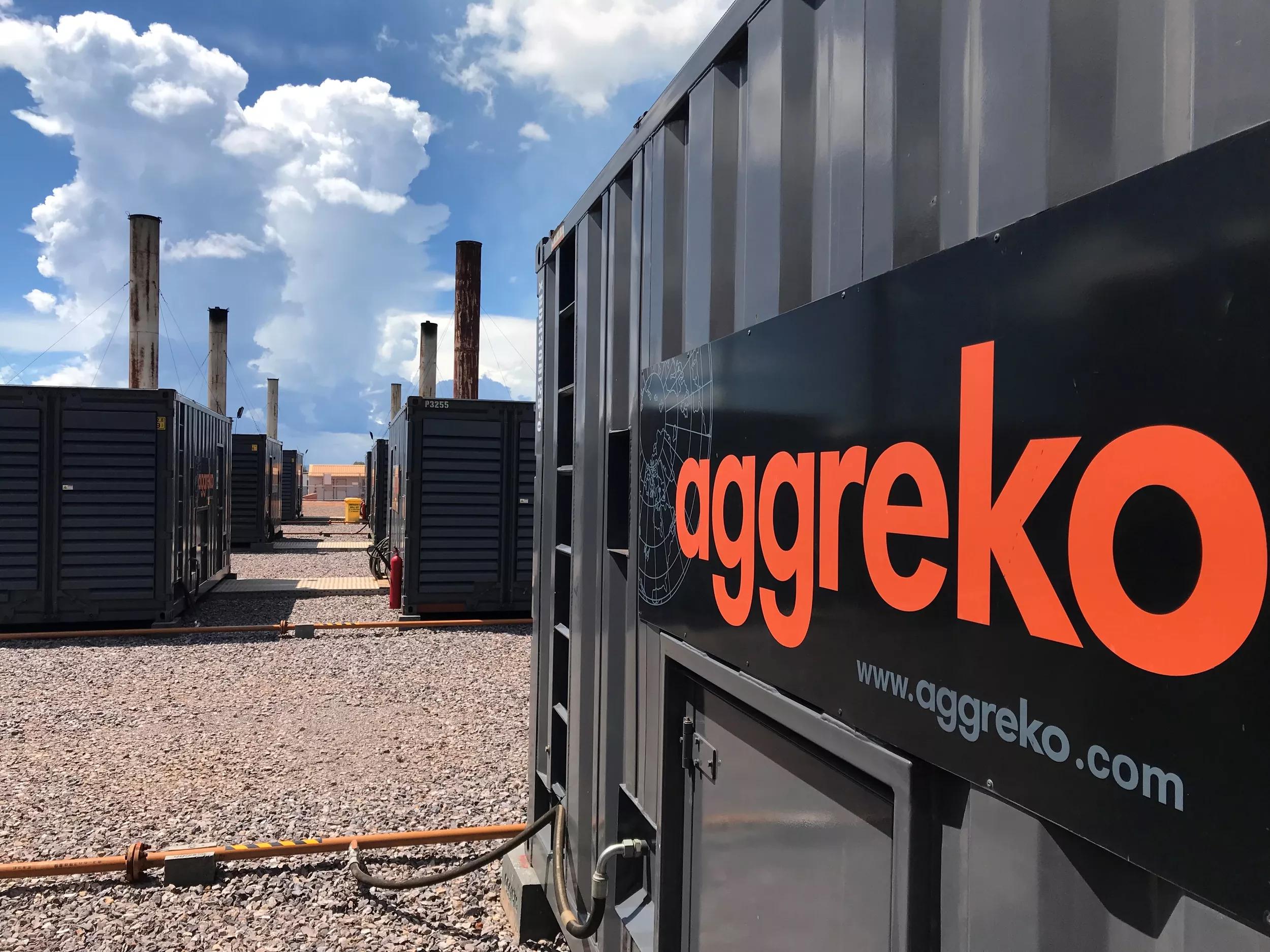 Generators with an Aggreko logo line a gravel pathway under a blue sky with clouds, showcasing industrial equipment.