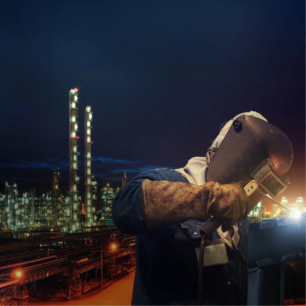 A worker with a safety mask welding at night and a petrochemical plant behind them