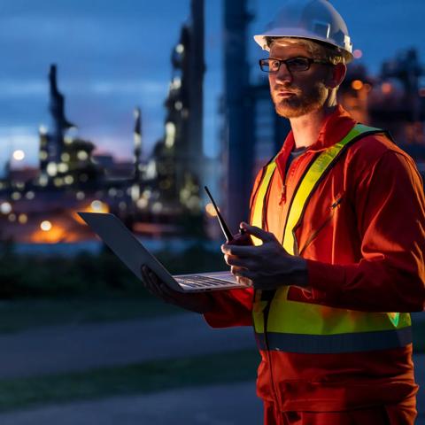 A worker holding a laptop and a cellphone at night, a power plant blurred in the background