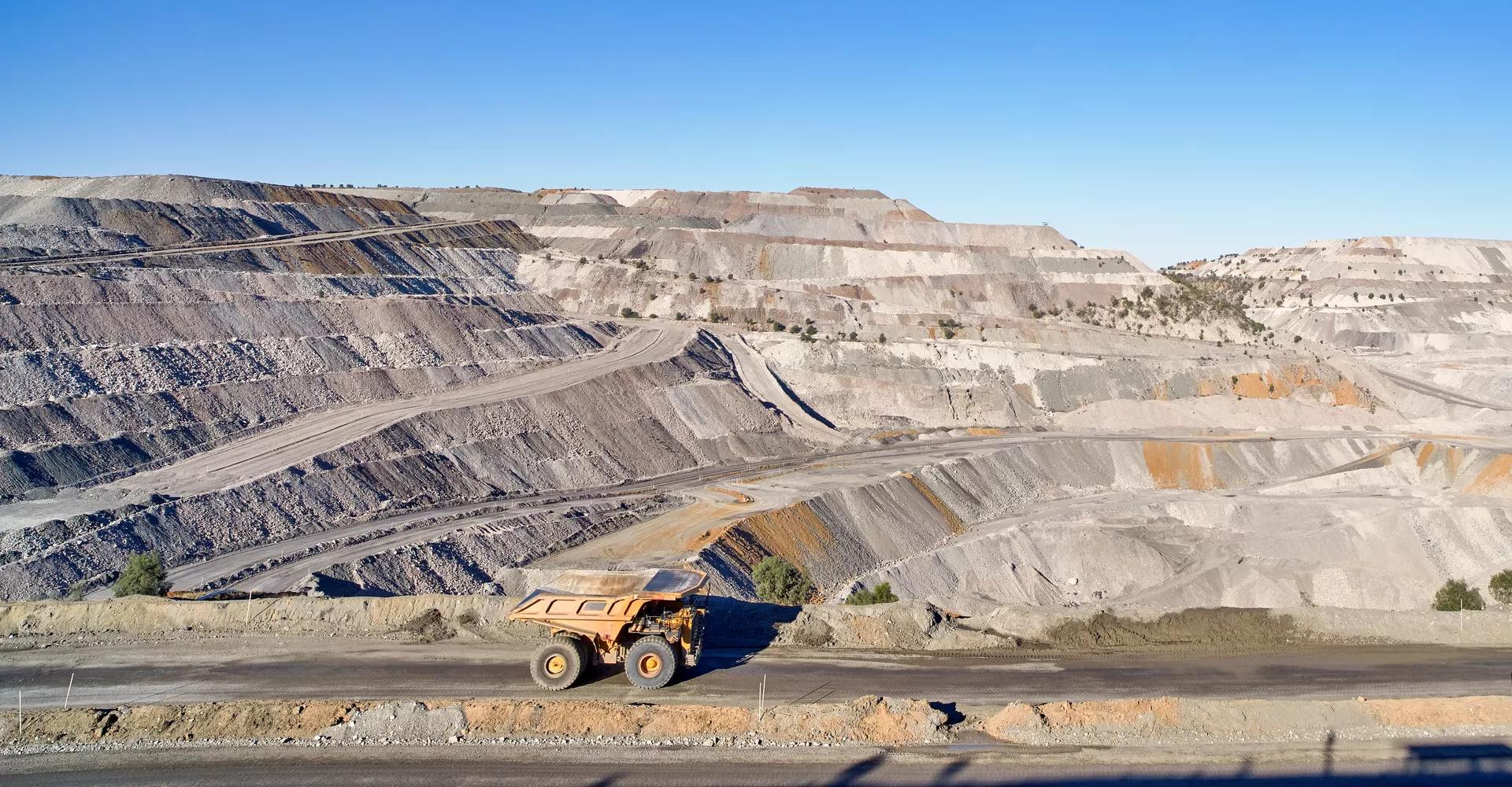 A wide panoramic view of a massive, terraced open-pit mine or quarry with light gray and brown layers of earth under a clear blue sky.
