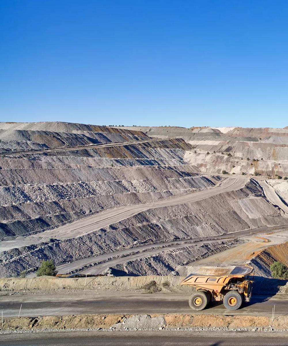A wide panoramic view of a massive, terraced open-pit mine or quarry with light gray and brown layers of earth under a clear blue sky.
